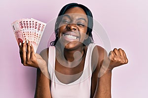 Young african american woman holding 100 new taiwan dollars banknotes screaming proud, celebrating victory and success very