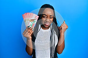 Young african american woman holding 100 new zealand dollars banknote screaming proud, celebrating victory and success very