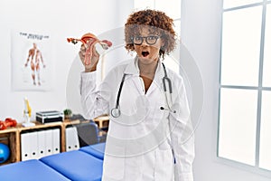 Young african american woman holding anatomical model of female genital organ scared and amazed with open mouth for surprise,