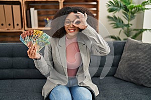 Young african american woman holding australian dollars at consultation office smiling happy doing ok sign with hand on eye