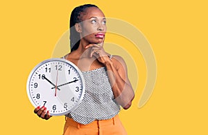 Young african american woman holding big clock serious face thinking about question with hand on chin, thoughtful about confusing