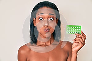 Young african american woman holding birth control pills puffing cheeks with funny face