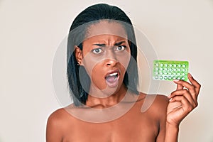 Young african american woman holding birth control pills in shock face, looking skeptical and sarcastic, surprised with open mouth