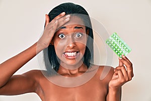Young african american woman holding birth control pills stressed and frustrated with hand on head, surprised and angry face