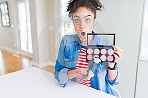 Young african american woman holding cosmetics blush palette scared in shock with a surprise face, afraid and excited with fear