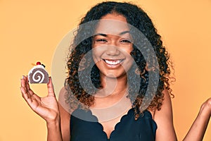 Young african american woman holding cupcake screaming proud, celebrating victory and success very excited with raised arm