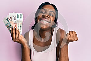 Young african american woman holding czech koruna banknotes screaming proud, celebrating victory and success very excited with