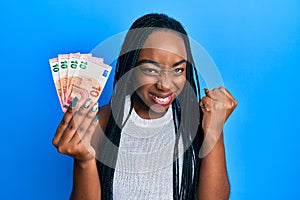 Young african american woman holding euro banknotes screaming proud, celebrating victory and success very excited with raised arm