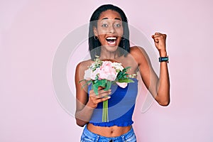 Young african american woman holding flowers screaming proud, celebrating victory and success very excited with raised arms