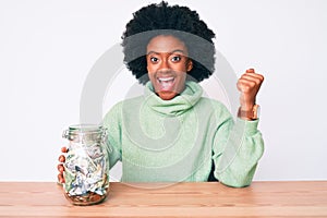 Young african american woman holding jar with savings screaming proud, celebrating victory and success very excited with raised