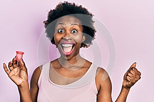 Young african american woman holding menstrual cup screaming proud, celebrating victory and success very excited with raised arms