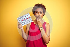 Young african american woman holding menstruation calendar over isolated yellow background cover mouth with hand shocked with