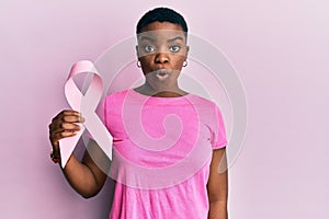 Young african american woman holding pink cancer ribbon scared and amazed with open mouth for surprise, disbelief face