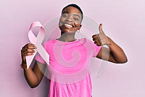 Young african american woman holding pink cancer ribbon smiling happy and positive, thumb up doing excellent and approval sign