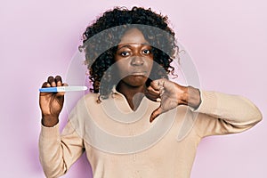 Young african american woman holding pregnancy test result with angry face, negative sign showing dislike with thumbs down,