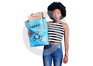 Young african american woman holding recycling bag with paper and cardboard scared and amazed with open mouth for surprise,