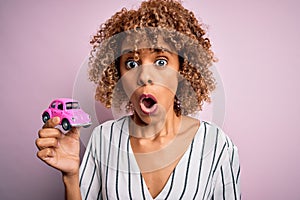 Young african american woman holding small pink car standing over isolated pink background scared in shock with a surprise face,