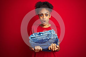 Young african american woman holding stack of jeans over isolated red background scared in shock with a surprise face, afraid and