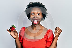 Young african american woman holding strawberry screaming proud, celebrating victory and success very excited with raised arms