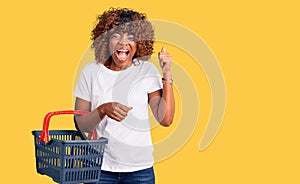 Young african american woman holding supermarket shopping basket screaming proud, celebrating victory and success very excited