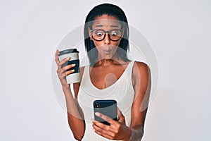 Young african american woman holding takeaway cup of coffee using smartphone making fish face with mouth and squinting eyes, crazy