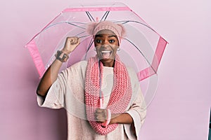 Young african american woman holding umbrella screaming proud, celebrating victory and success very excited with raised arms