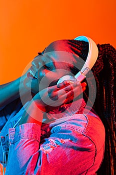 Young african-american woman listening to music in headphones in neon light. Female portrait. Concept of human emotions