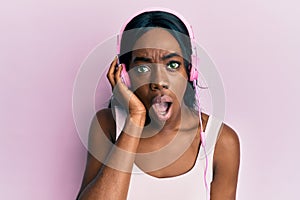 Young african american woman listening to music using headphones scared and amazed with open mouth for surprise, disbelief face