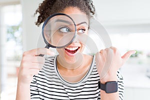 Young african american woman looking through magnifying glass pointing and showing with thumb up to the side with happy face