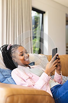 A young African American woman lounges on a sofa, smiling at her phone, with copy space