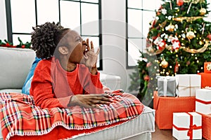 Young african american woman lying on the sofa by christmas tree shouting and screaming loud to side with hand on mouth