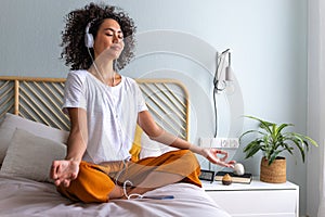 Young woman meditating at home sitting on bed following online meditation, listening with headphones, using phone app.