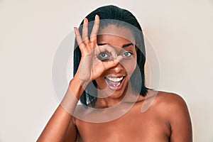 Young african american woman naked over background smiling happy doing ok sign with hand on eye looking through fingers