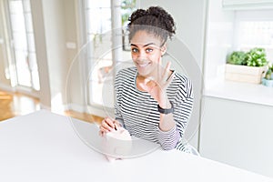 Young african american woman putting a coin inside piggy bank as invest or saving doing ok sign with fingers, excellent symbol