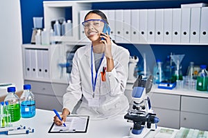 Young african american woman scientist talking on the smartphone write on report document at laboratory