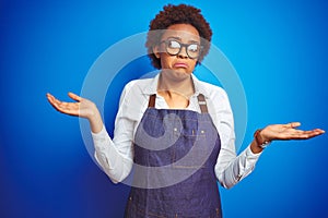 Young african american woman shop owner wearing business apron over blue background clueless and confused expression with arms and