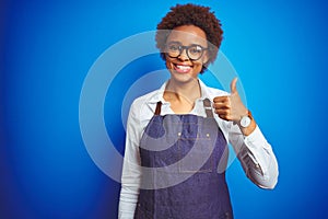 Young african american woman shop owner wearing business apron over blue background doing happy thumbs up gesture with hand