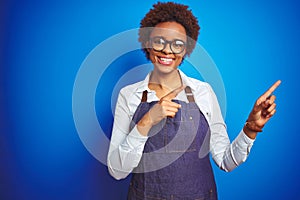 Young african american woman shop owner wearing business apron over blue background smiling and looking at the camera pointing