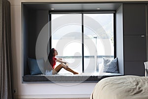 A young African American woman sits by a large window, gazing outside with copy space