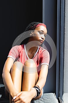 A young African American woman sits by a window, sunlight highlighting her thoughtful expression
