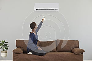 Young African American woman sitting on couch at home switches air conditioner on wall