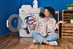 Young african american woman sitting on the floor doing laundry clueless and confused expression with arms and hands raised