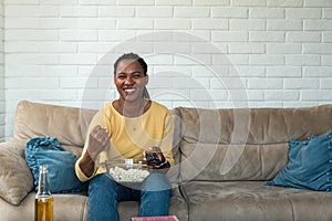 Young African American woman sitting at home on sofa watching sport game on TV, eating popcorn and cheering. Mother enjoying time