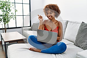 Young african american woman sitting on the sofa at home using laptop with a big smile on face, pointing with hand and finger to