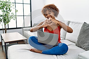 Young african american woman sitting on the sofa at home using laptop smiling in love doing heart symbol shape with hands