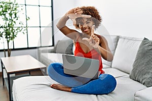 Young african american woman sitting on the sofa at home using laptop smiling making frame with hands and fingers with happy face