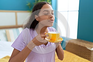 Young african american woman smelling cup of coffee sitting on bed at bedroom