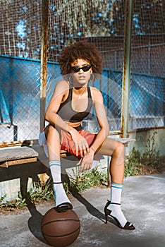 Young african-american woman in sportive attire and high heels sitting on bench with basketball ball under