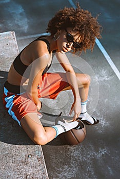 Young african-american woman in sportive attire and high heels sitting on bench with basketball ball under