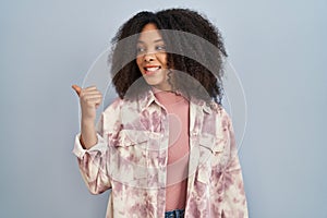 Young african american woman standing over blue background smiling with happy face looking and pointing to the side with thumb up